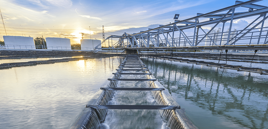 A water treatment plant with a water channel flowing into a large water tank. The sun is setting behind the plant, casting a warm glow.