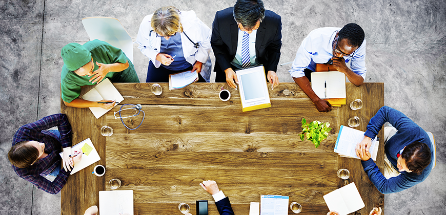 A top-down view of a diverse group of people, including doctors, nurses, and business professionals, sitting around a long wooden table.