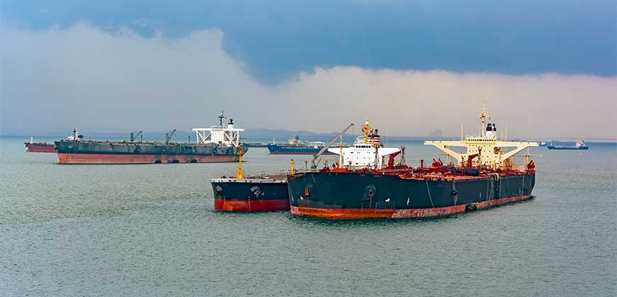 A group of massive oil tankers, some with cranes, are moored in calm waters. A dramatic storm cloud looms in the distance.