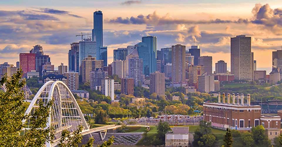 A panoramic view of the Edmonton skyline at sunset, featuring tall skyscrapers, a bridge, and green spaces.