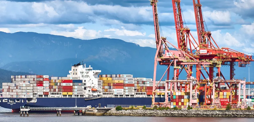 A large cargo ship with colorful shipping containers is docked at a port. In the background, there are large red cranes and more containers stacked on the dock. Mountains are visible in the distance.