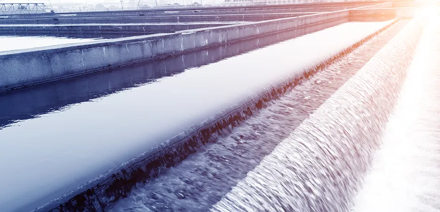 A close-up view of water flowing through a series of channels in a water treatment plant. The water is a dark blue colour, and the channels are lined with concrete.