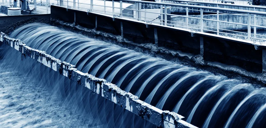 A close-up view of water flowing through a series of channels in a water treatment plant. The water is a dark blue color, and the channels are lined with concrete.