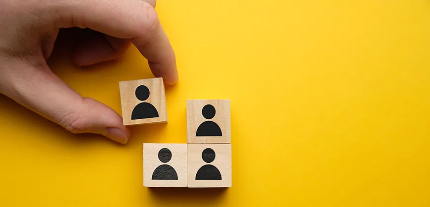 A hand placing a wooden block with a person icon on top of a stack of three similar blocks, arranged in a pyramid shape on a yellow background.