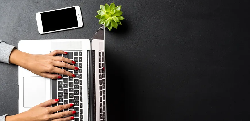 A person's hands typing on a laptop keyboard, with a phone and a small plant on a black desk.