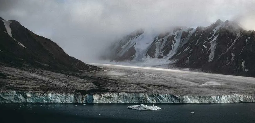 A vast, icy glacier extends from snow-capped mountains into a calm, dark body of water. A small iceberg floats nearby.