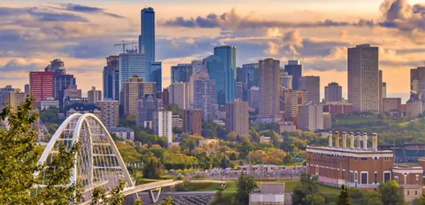 A panoramic view of the Edmonton skyline at sunset, featuring tall skyscrapers, a bridge, and green spaces.